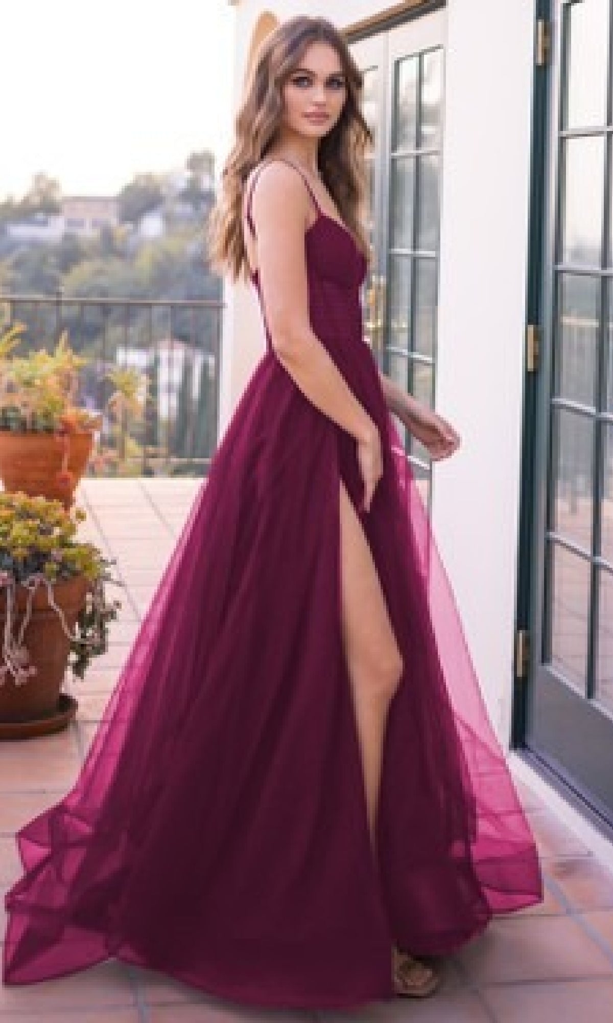A woman in the Yasmin by Velvi Sheer-Corset Long Prom Ball Gown, an affordable sleeveless floor-length dress with a high slit, stands on a patio near potted plants and glass doors.